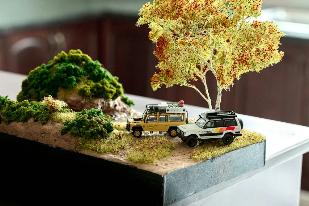 A miniature landscape with a small tree, moss and two toy off-road cars.